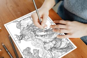 A person's hands holding colored pencils, carefully coloring a detailed western-themed coloring page. The page features a cowboy on horseback riding beside a long-horn cow, set against a rustic, open landscape.