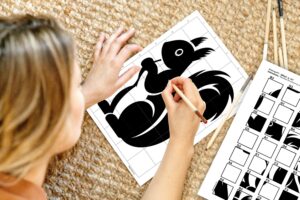 A woman holding a pencil and finishing a mystery grid drawing puzzle of a squirrel. The completed drawing is visible above, with the corresponding grid template next to it.