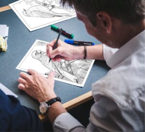 A man coloring two detailed coloring pages. One page depicts a robin perched on a branch, while the other features a small parrot in its natural habitat, surrounded by foliage. The man is using colored pencils to bring the birds to life.