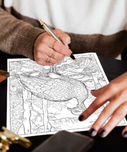 A person’s hand holding a pen, coloring a detailed coloring page of a wild turkey in its natural habitat. The page showcases the turkey in a wooded environment, surrounded by trees and foliage.