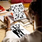 A woman holding up a completed mystery grid drawing template of an African elephant, with a pencil in hand, sitting in front of a grid sheet where she has recreated the drawing which she has clipped onto a clipboard.