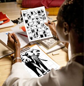 A woman holding up a completed mystery grid drawing template of an African elephant, with a pencil in hand, sitting in front of a grid sheet where she has recreated the drawing which she has clipped onto a clipboard.