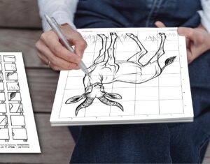 A man using a pen to complete a mystery grid drawing puzzle of an African klipspringer, following the grid instructions and copying squares from the template on a bench next to him.