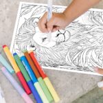 A person's hand holding a marker, preparing to color a detailed coloring page of an African lion with a lush, abundant mane.