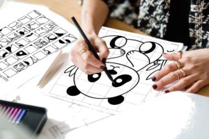 A woman’s hands drawing on a mystery grid printable worksheet, revealing a simple panda bear surrounded by leaves. The grid is completely filled with pencil marks.